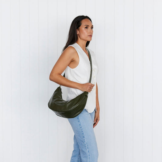 A woman walking away from the camera, wearing a white top and blue jeans, carrying a green slouchy shoulder bag with a zipper.