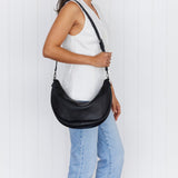 A person wearing a white tank top and blue jeans is shown from the back, carrying a black crossbody bag over shoulder on long strap.