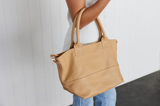 A woman holding a beige leather tote bag over their forearm against a white background.