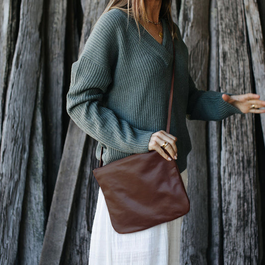 A person wearing a green sweater and a white skirt is carrying a brown leather crossbody bag.