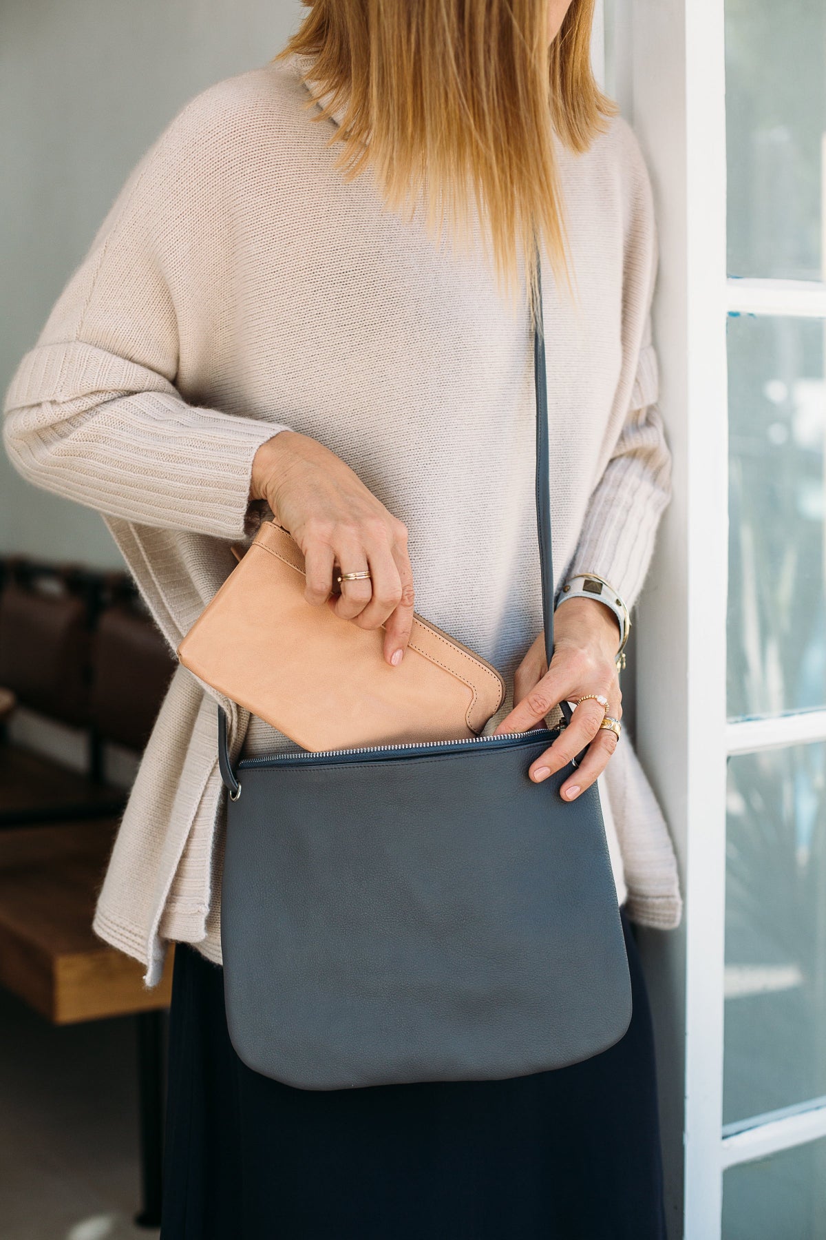 Person holding a grey crossbody bag with a blush wallet being placed inside, standing doorway.