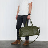 A man wearing a white t-shirt, dark jeans, and brown boots stands in a studio holding a large army
 green leather duffel bag.
