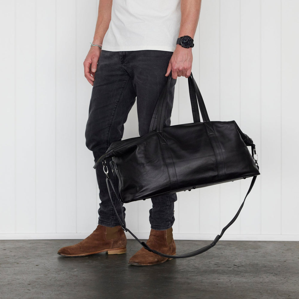 Black leather weekender / duffle bag handheld by man in black jeans and brown boots
