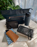 A collection of leather bags and accessories arranged on a white textured surface including a tote bag, a water bottle, a pouch, sunglasses holder and a wallet.