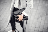 Woman in black jeans with grey coat holding a black bag with strap haging down